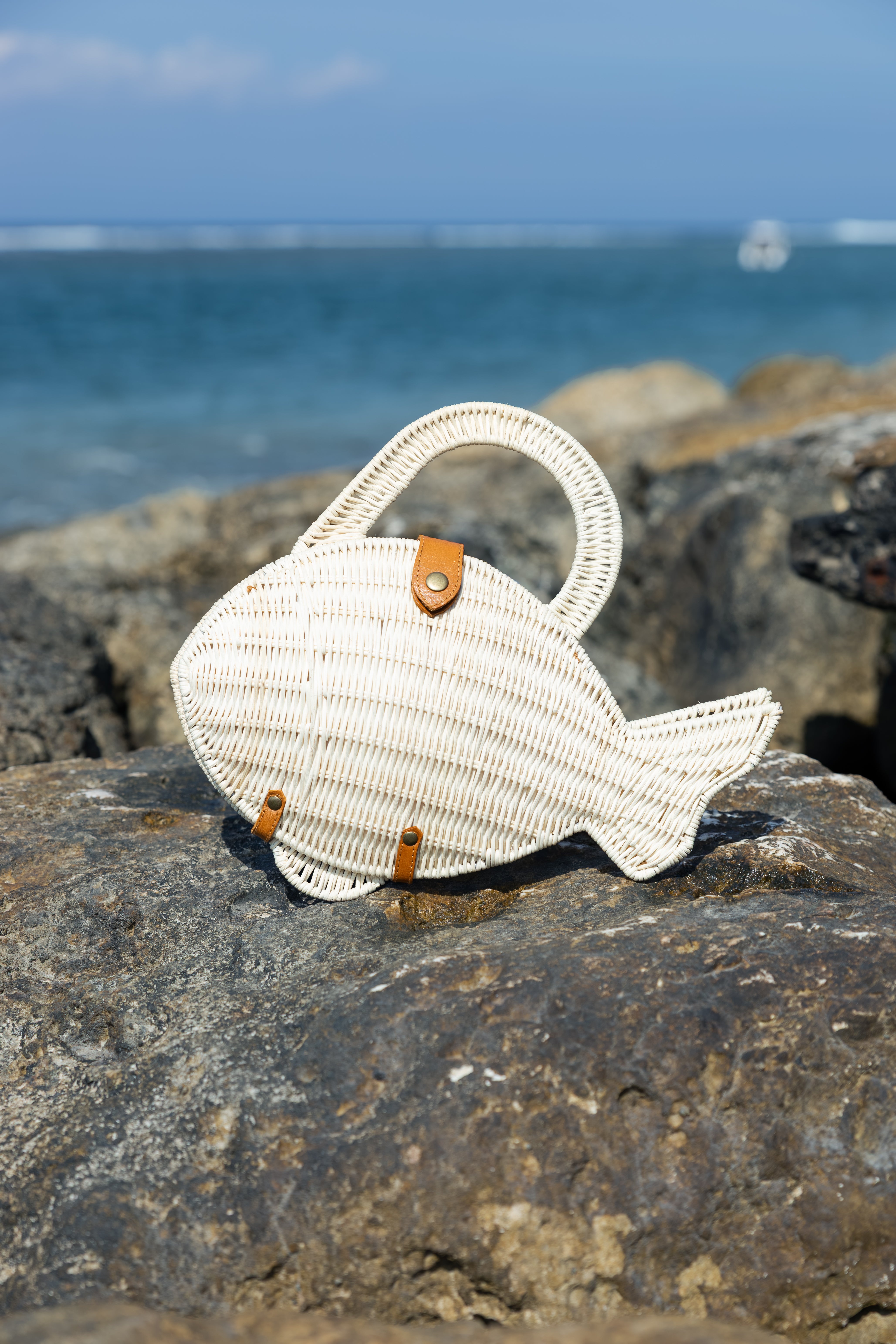 A natural fish-shaped handwoven straw handbag with a top handle and small brown leather accents, resting on a large rock with the ocean and sky blurred in the background.