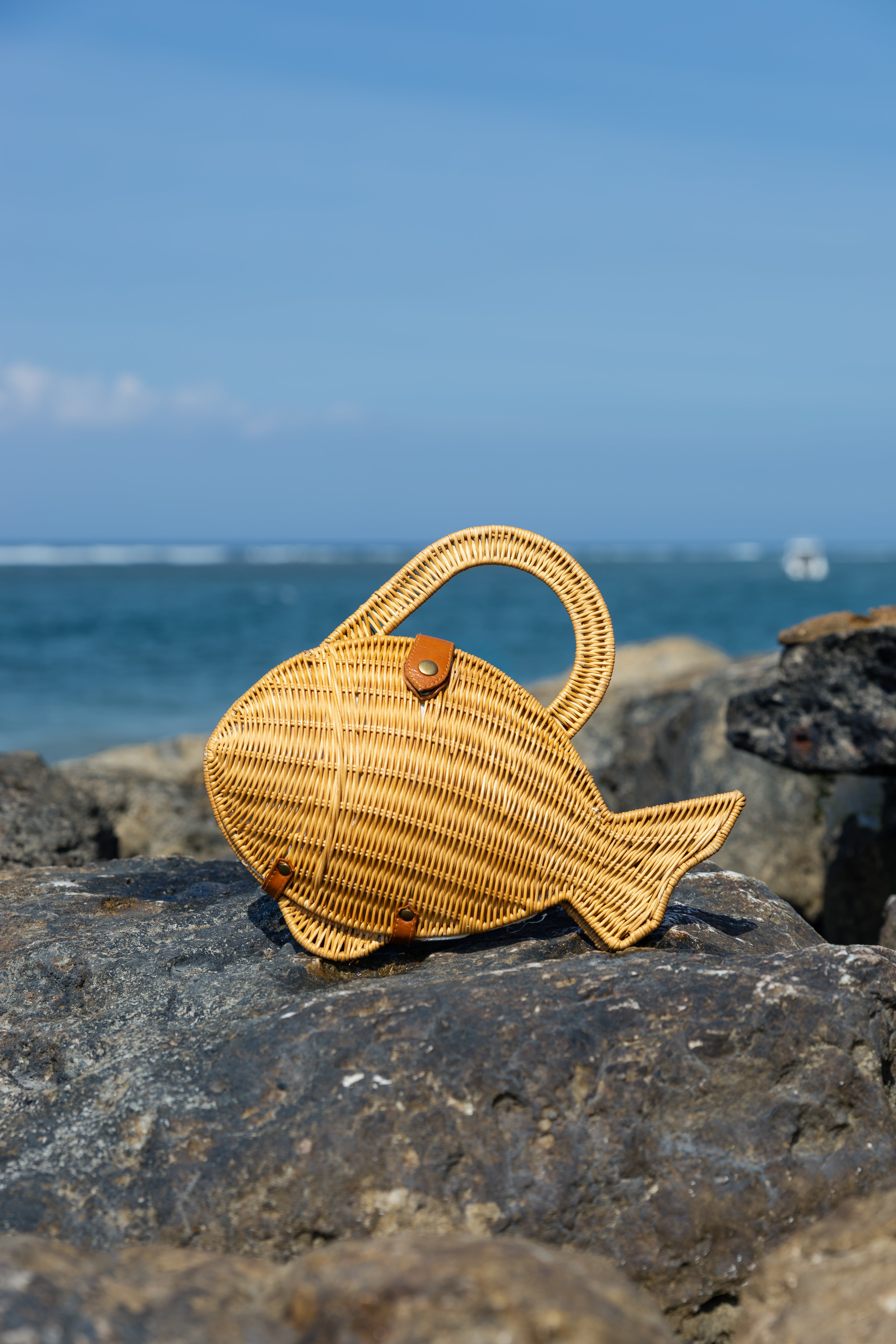 A woven fish-shaped handbag made of natural straw or wicker, featuring a top handle and a small brown button closure, resting on a dark rock with a blurred ocean and blue sky background.