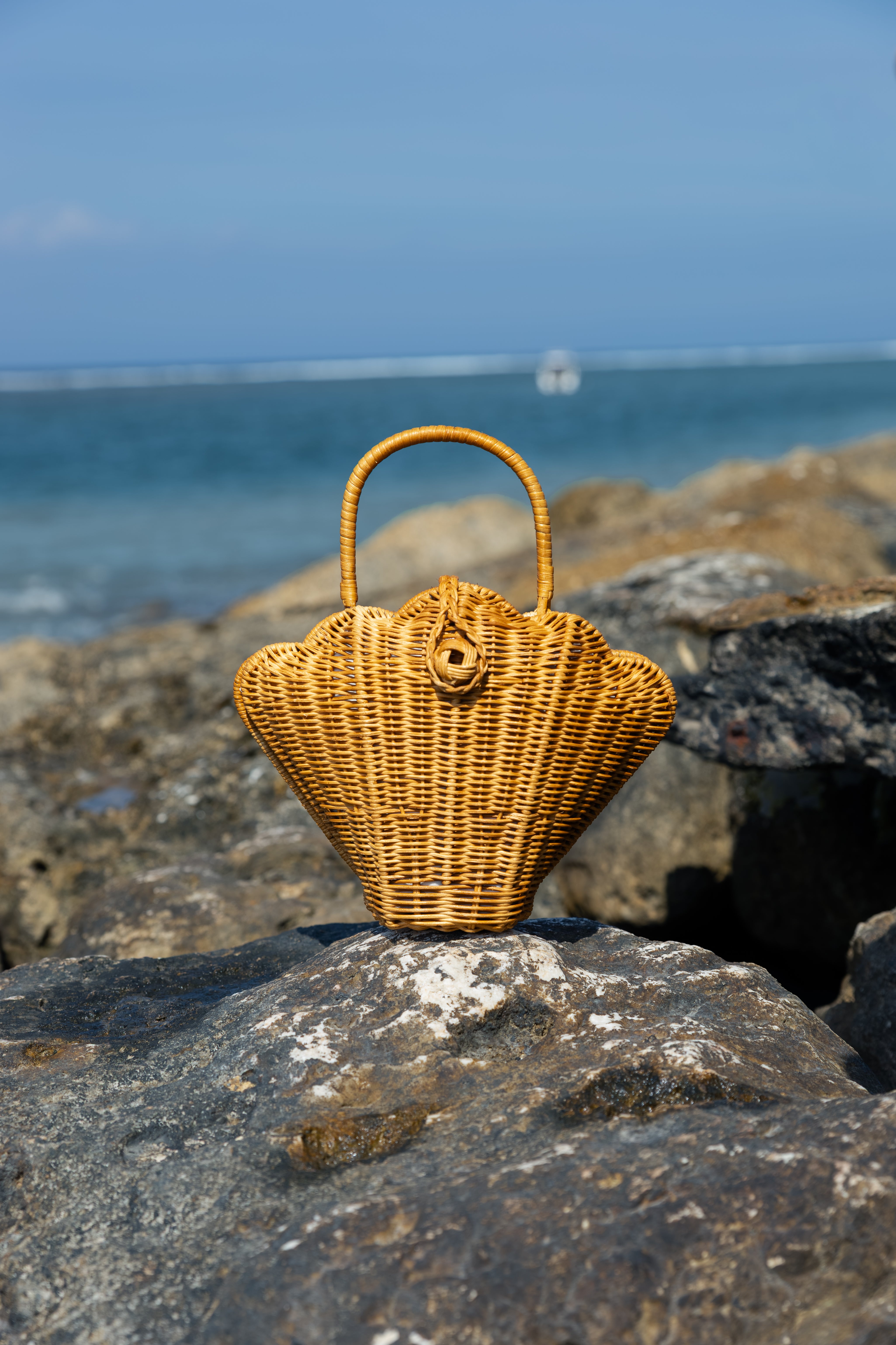 A natural wicker sea shell-shaped handbag with a curved top handle, styled on a dark rocky shore with the ocean and blue sky blurred in the background.