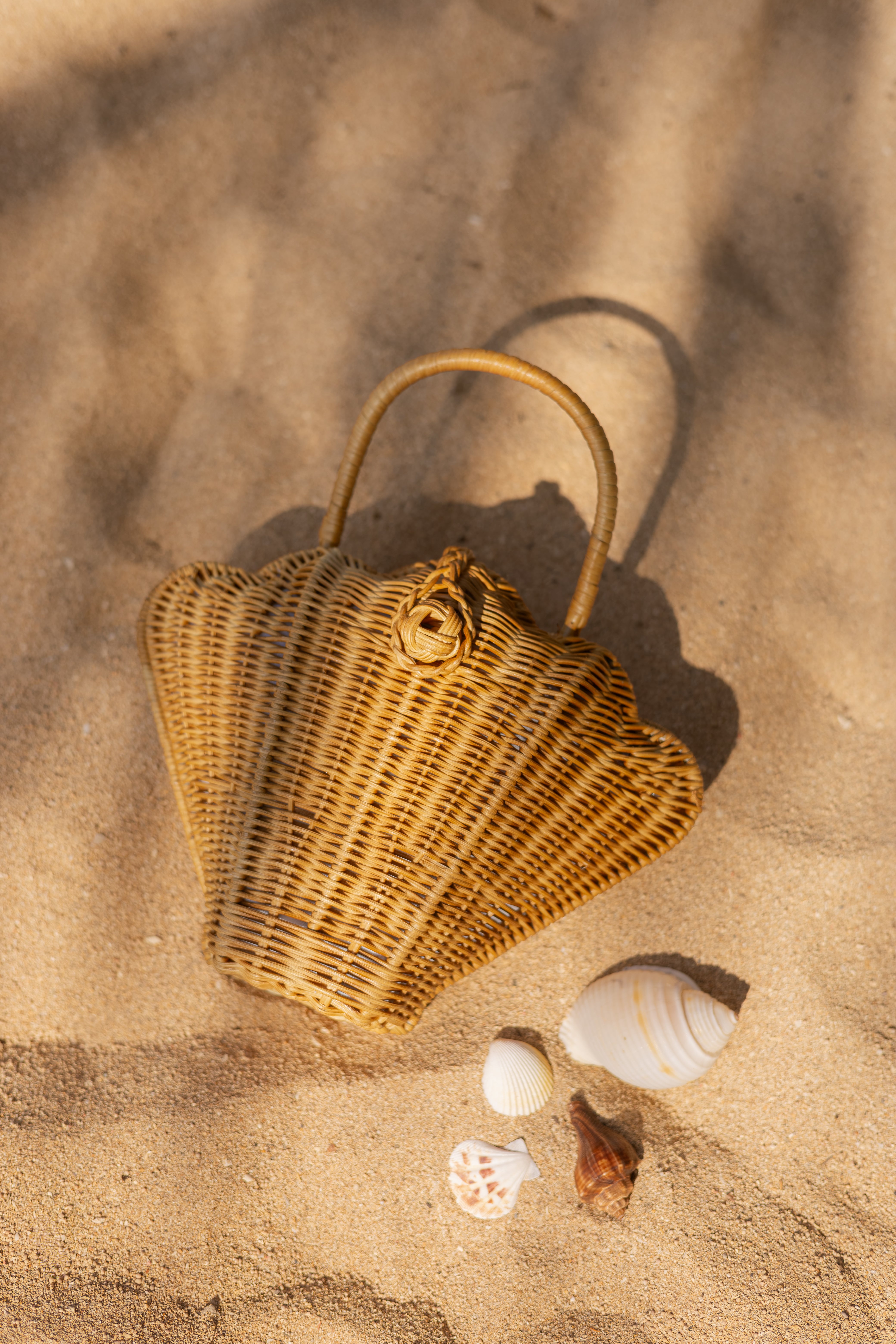 A woven sculptural seashell-shaped handbag with a curved top handle, styled on sand.