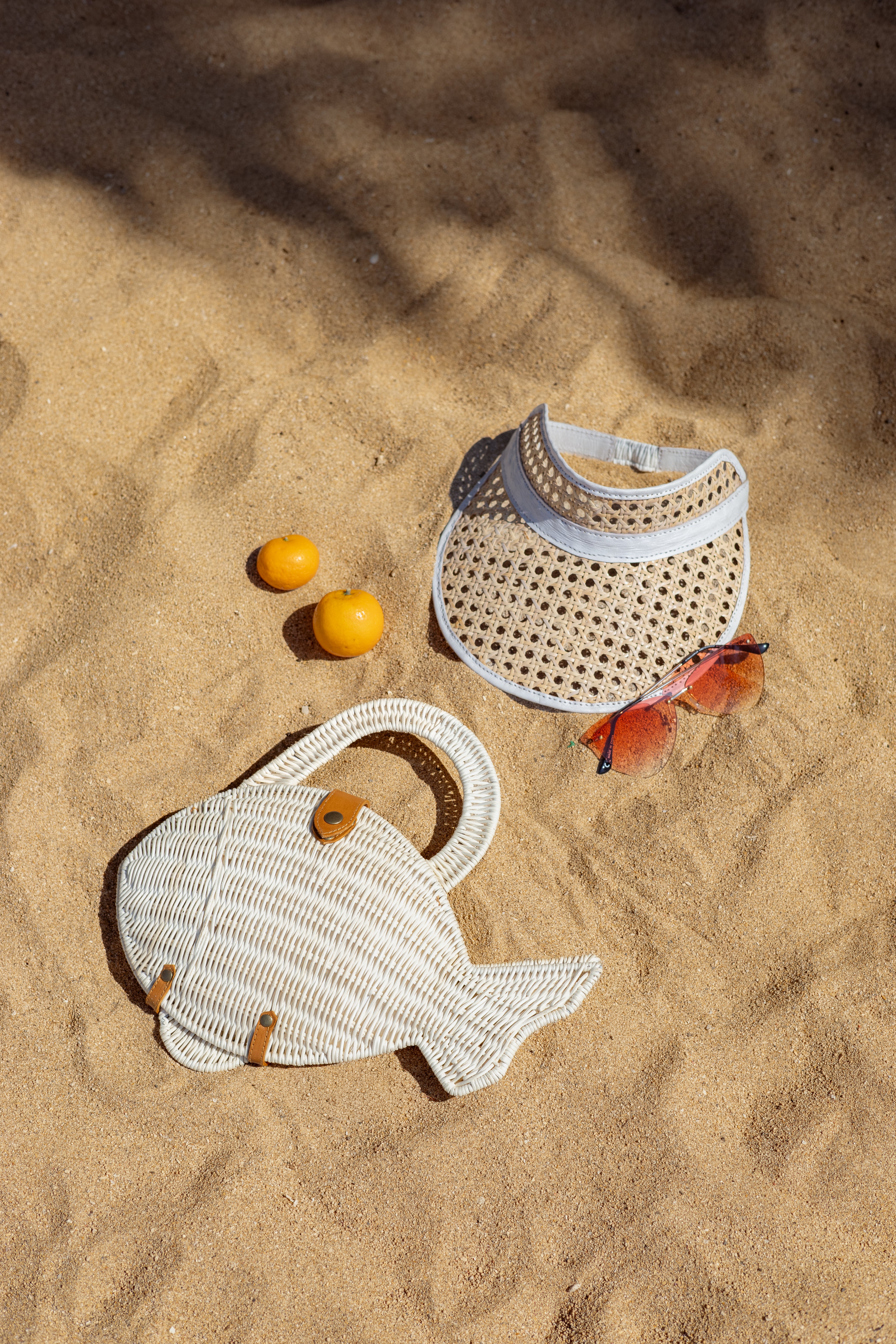 Handwoven fish-shaped handbag in natural rattan  lies on sandy beach, styled with a cane webbing rattan and leather trim visor hat, pinkish sunglasses, and two small oranges. The scene is photographed from above in bright sunlight.